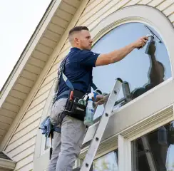 Professional window cleaner on a ladder using a squeegee to clean an exterior arched window, Colorado Springs, CO.